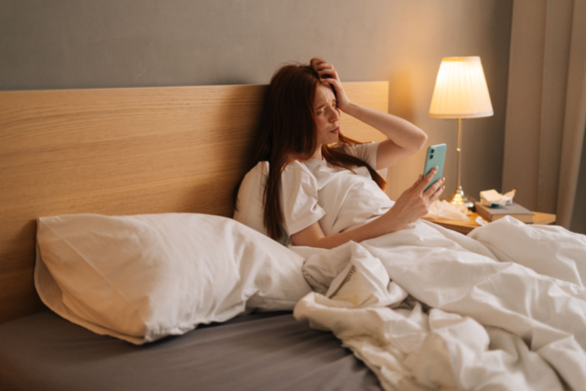 Överväger nattlig melatoninanvändning - kvinna i säng med telefon ser trött ut Kvinna i säng ser trött ut medan hon använder telefon, möjligen reflekterande över nattlig melatoninanvändning